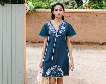 May include: A woman wearing a blue denim dress with white embroidered flowers. She is carrying a white and yellow bag and wearing white sneakers.