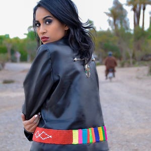 May include: A woman wearing a black leather jacket with a colourful woven belt. The belt has a red base with white stitching and a multi-coloured stripe pattern.