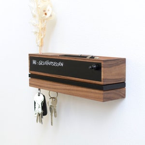 Op de afbeelding: Een houten sleutelhanger met een zwart schoolbord aan de bovenkant. Op het schoolbord staat de tekst "MI: GESCHÄFTSESSEN". Er hangen sleutels aan de onderkant van de hanger.