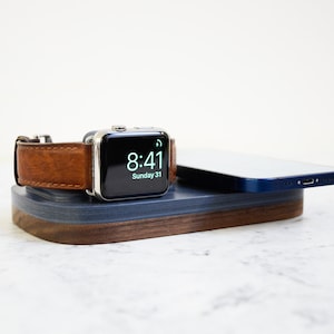 Könnte beinhalten: Eine Ladestation aus Holz mit blauer und brauner Oberfläche. Eine silberne Apple Watch mit einem braunen Lederarmband befindet sich auf der linken Seite der Station. Ein blaues Smartphone befindet sich auf der rechten Seite der Station. Das Zifferblatt der Uhr zeigt die Zeit 8:41 und das Datum Sonntag 31.
