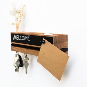 Op de afbeelding: Houten sleutelhouder met een schoolbord met de tekst "WELCOME" in het wit. De sleutelhouder heeft een zwarte accentstreep en een kleine sleuf voor post of notities. Sleutels hangen aan de onderkant en een kleine droogbloemarrangement staat bovenop.