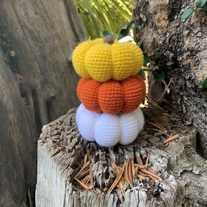 May include: A stack of three crocheted pumpkins in yellow, orange, and white. The pumpkins are sitting on a tree stump.