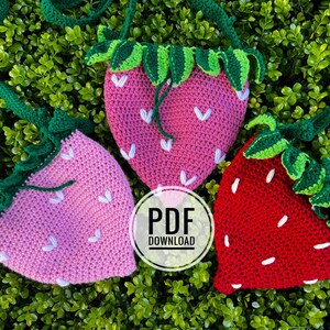 May include: Three crocheted strawberry-shaped bags with green leaves. The bags are pink, red, and pink. The bags have white stitching and are on a bed of green leaves. The text "PDF DOWNLOAD" is in a white circle in the center of the image.