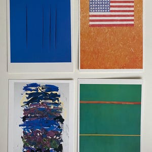 May include: Four framed art prints, each with a different abstract design. The first print features a solid blue background with four vertical white lines. The second print features a red, white, and blue American flag on a textured orange background. The third print features a colorful abstract design with blue, purple, and yellow paint strokes. The fourth print features a green background with two horizontal lines, one orange and one yellow.