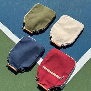 May include: Four rectangular canvas pouches in olive green, cream, navy blue, and red. Each pouch features a light-colored zipper closure, a brown leather-like trim at the base, and a small metal D-ring attachment. The red pouch includes an extra zippered pocket.