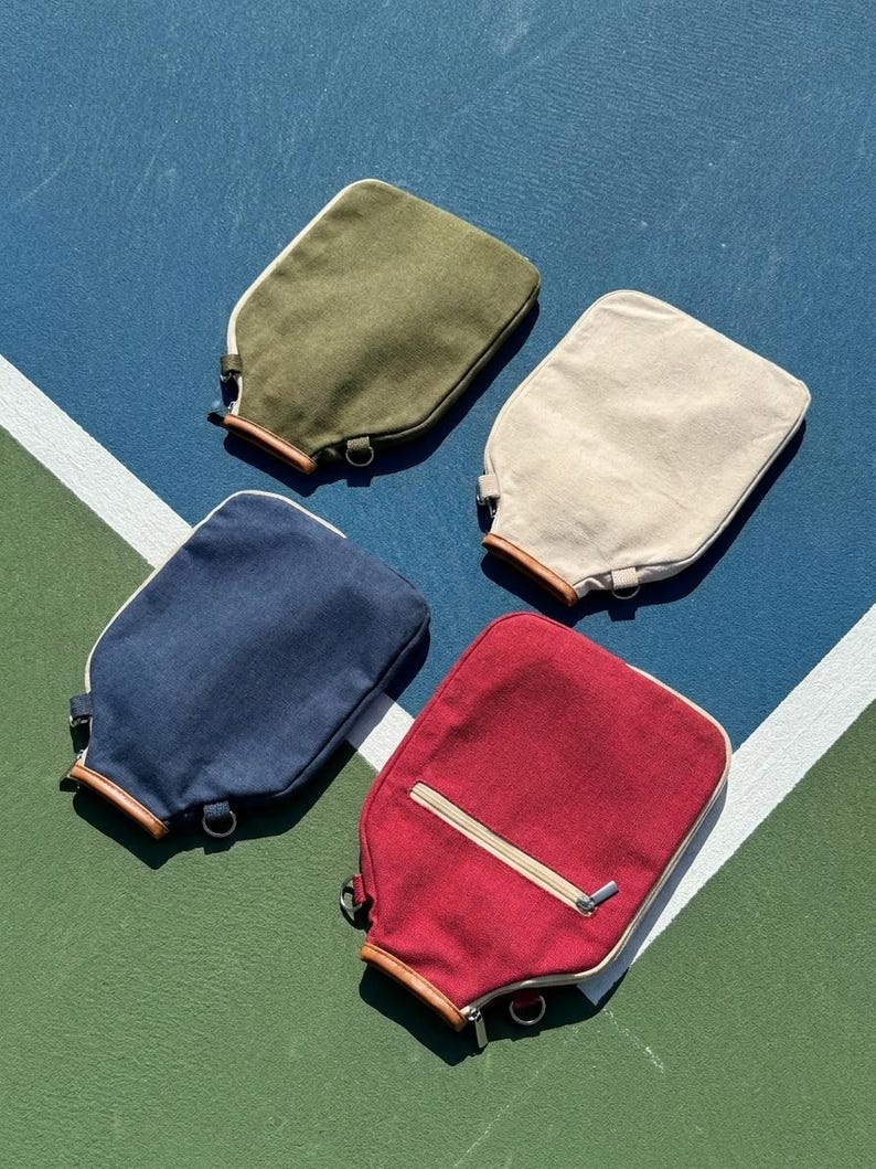 May include: Four rectangular canvas pouches in olive green, cream, navy blue, and red. Each pouch features a light-colored zipper closure, a brown leather-like trim at the base, and a small metal D-ring attachment. The red pouch includes an extra zippered pocket.
