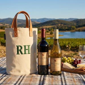 Puede incluir: Una escena de picnic con una bolsa de vino de lona con asas de cuero marrón y las letras bordadas "RH". Dos botellas de vino, una roja y otra blanca, se encuentran junto a una tabla de madera con queso, galletas y uvas. También hay dos copas de vino.