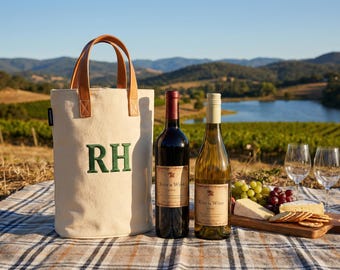 May include: A picnic scene with a canvas wine tote with brown leather handles and the embroidered letters "RH". Two wine bottles, a red and a white, sit next to a wooden board with cheese, crackers, and grapes. Two wine glasses are also present.