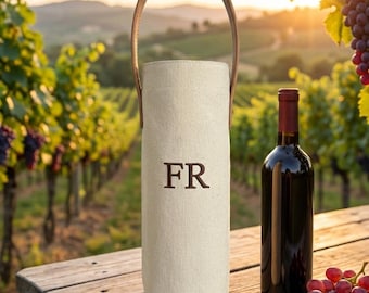May include: A beige wine bottle tote with a brown leather handle, featuring the embroidered letters "FR". A bottle of red wine and a bunch of grapes sit on a wooden table. The background shows a vineyard.