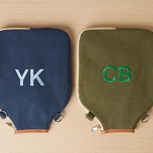 May include: Two rectangular canvas bags with rounded top corners, one in navy blue with light blue embroidered initials YK and one in olive green with green embroidered initials CB. Both feature a zipper closure, metal hardware, and brown trim.