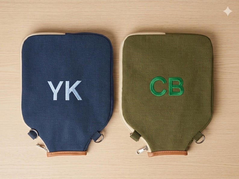 May include: Two rectangular canvas bags with rounded top corners, one in navy blue with light blue embroidered initials YK and one in olive green with green embroidered initials CB. Both feature a zipper closure, metal hardware, and brown trim.