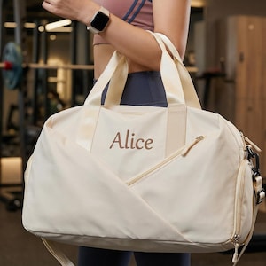 Könnte beinhalten: Cremefarbene Sporttasche mit dem Namen "Alice" in Braun gestickt. Die Tasche hat eine Reißverschluss-Vordertasche, einen Schultergurt und zwei obere Griffe. Die Tasche ist für den Gebrauch im Fitnessstudio konzipiert.