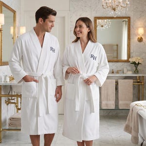 May include: Two people wearing white bathrobes with the embroidered initials "RH" in navy blue. The robes have a textured, ribbed design and are tied at the waist with a matching belt. The setting is a luxurious bathroom.