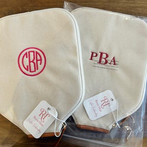 May include: Two off-white canvas pickleball paddle covers with brown leather trim at the base. One cover features a circular hot pink monogram with the letters CBA, while the other has red embroidered letters PBA. Each has a Rosa Hue brand tag.