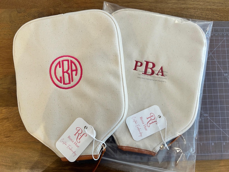 May include: Two off-white canvas pickleball paddle covers with brown leather trim at the base. One cover features a circular hot pink monogram with the letters CBA, while the other has red embroidered letters PBA. Each has a Rosa Hue brand tag.