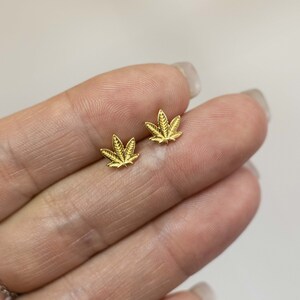 Puede incluir: Un par de pendientes de botón de hoja de marihuana de color dorado.
