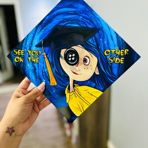 Pode incluir: Um chap&eacute;u de formatura decorado com o tema Coraline. O design mostra Coraline com um olho de bot&atilde;o, usando um capelo, contra um fundo azul rodopiante. O texto diz "See You on the Other Side."