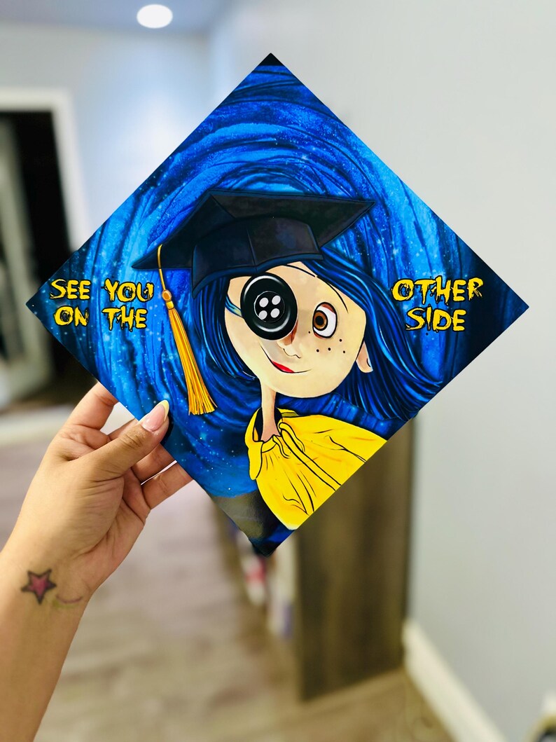 Pode incluir: Um chap&eacute;u de formatura decorado com o tema Coraline. O design mostra Coraline com um olho de bot&atilde;o, usando um capelo, contra um fundo azul rodopiante. O texto diz "See You on the Other Side."