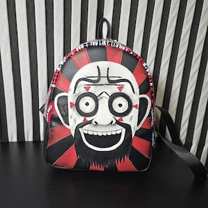 Puede incluir: Una mochila negra y roja con una cara de dibujos animados blanca en la parte delantera. La cara tiene una barba negra, ojos blancos y una amplia sonrisa. La mochila tiene una correa negra y un patrón de rayas negras y blancas alrededor del borde.