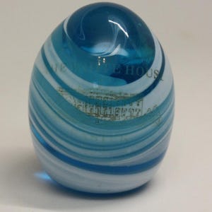 Puede incluir: Un pisapapeles de vidrio azul y blanco con forma de huevo y un diseño en espiral. El pisapapeles presenta una imagen de la Casa Blanca y el texto "WHITE HOUSE" es visible en la superficie.