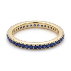 May include: A yellow gold eternity band featuring a continuous row of small, round, deep blue sapphires set along the outer circumference of the ring. The metal band has a polished finish and a consistent width throughout its circular design.