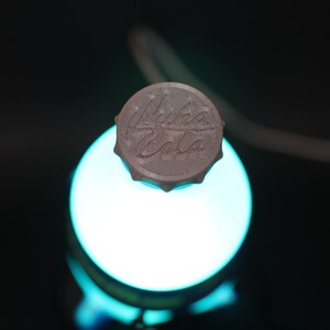 May include: A white bottle cap with the words "Nuka Cola" printed on it. The cap is sitting on top of a glowing blue light.