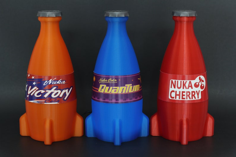 May include: Three rocket-shaped bottles of Nuka Cola, a fictional soda brand from the Fallout video game series. The bottles are orange, blue, and red, and each has a different flavor label: Victory, Quantum, and Cherry.