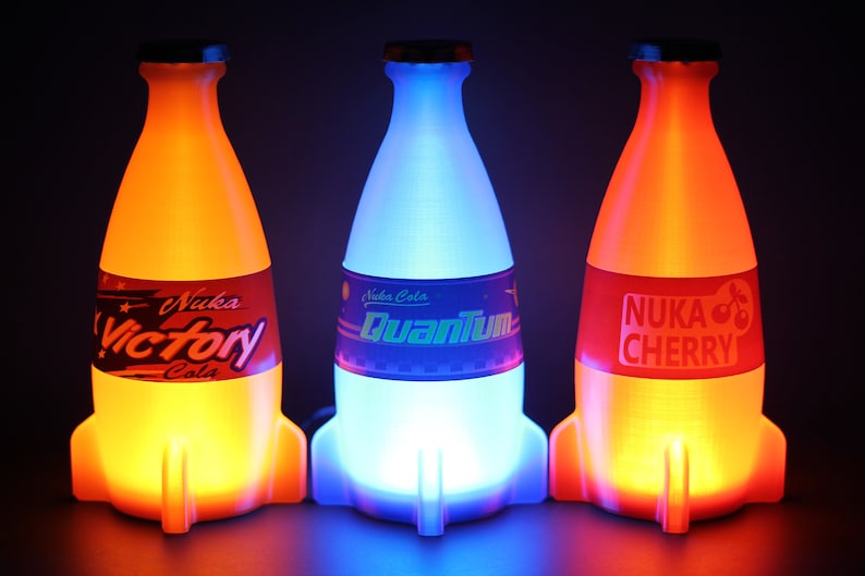 May include: Three glowing rocket-shaped bottles of Nuka Cola. The bottles are lit from within, with the left bottle glowing orange, the middle bottle glowing blue, and the right bottle glowing red. The bottles are labeled "Nuka Cola Victory", "Nuka Cola Quantum", and "Nuka Cola Cherry".