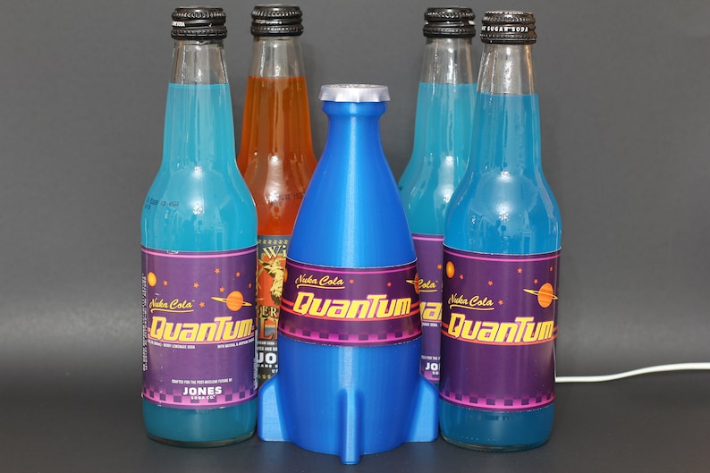May include: Five glass bottles of blue and orange soda with a rocket-shaped bottle stopper. The bottles are labeled "Nuka Cola Quantum" with a purple background and a retro-futuristic design.