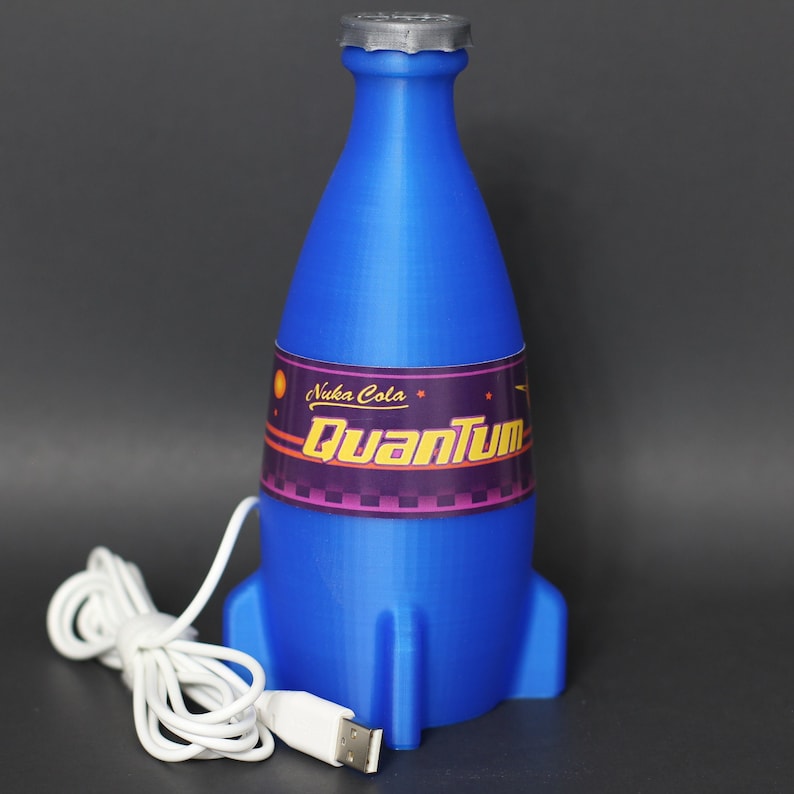 May include: A blue and white Nuka Cola Quantum bottle shaped like a rocket ship with a USB cable attached. The bottle has a label that says "Nuka Cola Quantum".
