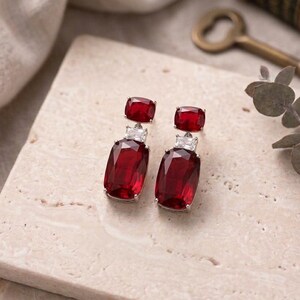 May include: A pair of silver earrings with a rectangular, deep red gemstone and a smaller red stone at the top. The earrings are displayed on a light-colored, textured surface, with a vintage key and book in the background.