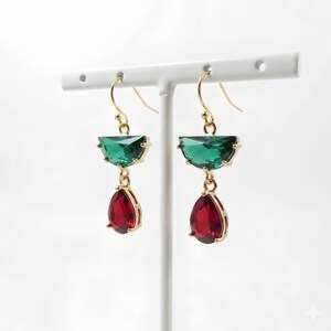 May include: A pair of gold-tone hook earrings featuring a faceted green semi-circle crystal suspended above a faceted red teardrop-shaped crystal. The earrings are displayed on a white T-shaped stand, showcasing the vibrant contrast of the gemstones.