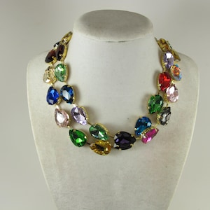 May include: A colorful statement necklace with teardrop-shaped gemstones in various colors, including blue, green, red, and purple. The gemstones are set in a gold-tone metal frame, creating a vibrant and eye-catching accessory. The necklace is displayed on a white mannequin.
