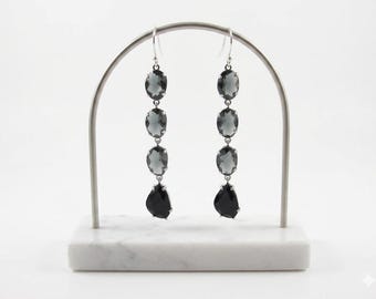 May include: Long drop earrings with silver hooks and settings. Each earring has three oval, smoky grey stones and a single, black teardrop-shaped stone at the bottom. The earrings are displayed on a white marble stand.