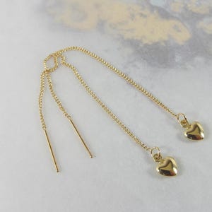 May include: Gold-colored threader earrings with heart charms. The earrings feature delicate chains that extend through the ear, with a small heart pendant dangling at the end. The earrings are displayed on a light-colored surface.