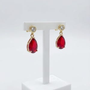 May include: A pair of gold-toned drop earrings featuring a clear, round, faceted stone at the top and a larger, teardrop-shaped, deep red faceted stone suspended below. The earrings are displayed on a white T-shaped stand against a plain background.