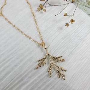 May include: A gold-colored necklace with a delicate, branch-like pendant. The pendant has a detailed, textured design. The necklace chain is thin and also gold-colored. The jewelry is displayed on a white, textured surface.