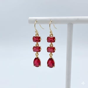 May include: A pair of gold-tone drop earrings featuring three red faceted stones each. The top two stones are rectangular, while the bottom stone is a teardrop shape. The earrings hang from gold-tone hooks and are displayed on a white stand.