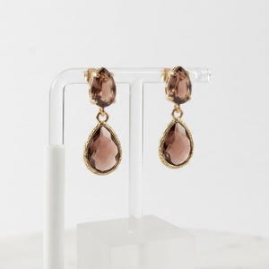 May include: A pair of gold-toned drop earrings featuring two faceted, pear-shaped brown crystals. The smaller crystal sits at the top, while the larger crystal hangs below, encased in a textured gold-toned metal frame with a secure post backing.