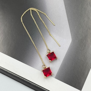 May include: A pair of gold chain earrings with red square-shaped gemstones. The earrings are dangling and have a simple design.