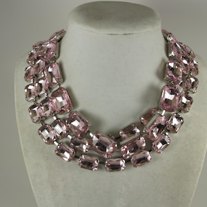 May include: A statement necklace featuring multiple rows of large, rectangular, light pink crystals. The crystals are set in a silver-tone metal and arranged in a layered design. The necklace is displayed on a mannequin bust.