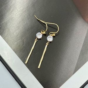 May include: A pair of gold dangle earrings with a small clear rhinestone at the top of each earring. The earrings have a thin, straight gold bar hanging from the rhinestone.