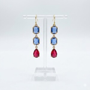 May include: A pair of gold-tone drop earrings featuring two stacked rectangular blue crystals followed by a single teardrop-shaped red crystal at the base. The earrings are suspended from gold-tone hooks and display a clear, faceted finish.