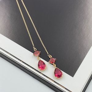 May include: Gold-coloured earrings with a chain design. Each earring has a square red gemstone and a teardrop-shaped red gemstone. The earrings are displayed on a black and white surface.