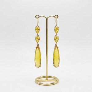 May include: A pair of long, yellow gemstone earrings on a gold-colored display stand. Each earring features three heart-shaped stones and a large teardrop-shaped stone. The earrings are displayed against a white background.