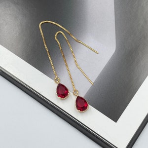 May include: A pair of gold dangle earrings with red teardrop-shaped crystals. The earrings have a delicate chain design and are perfect for adding a touch of sparkle to any outfit.