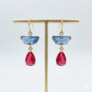 May include: A pair of gold-toned earrings. Each earring features a light blue, half-circle-shaped gem above a red, teardrop-shaped gem. The earrings are displayed on a gold stand against a white background.