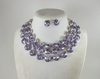 May include: A three-strand lavender crystal necklace and matching stud earrings set on a white mannequin bust. The necklace and earrings feature large, round, faceted crystals with a silver-tone metal setting.