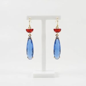 Puede incluir: Un par de pendientes colgantes dorados con un pequeño cristal rojo semicircular en la parte superior y un cristal azul más grande en forma de lágrima suspendido debajo. Los pendientes cuelgan de ganchos en un soporte de exhibición blanco.