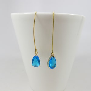 May include: A pair of gold-colored drop earrings with teardrop-shaped, bright blue gemstones. The earrings hang from thin, curved wires, and the gemstones are set in gold-colored bezels. The background is a white surface.